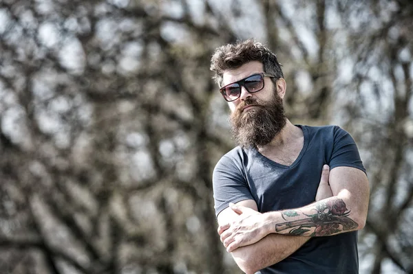 Homem barbudo sério de pé com os braços cruzados. Homem bonito com tatuagem vestindo óculos de sol na moda posando ao ar livre — Fotografia de Stock