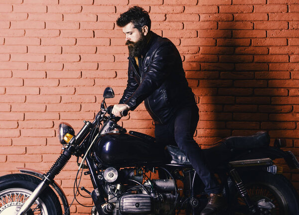 brutal biker on serious face in leather jacket gets on motorcycle. Man with beard, biker in leather jacket near motor bike in garage, brick wall background. Start of journey concept