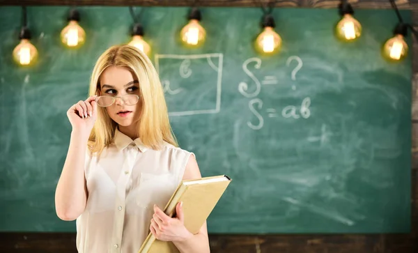 La mujer con libro comienza la lección, mira al público mientras se quita las gafas. El profesor se ve confiado en las gafas, de pie en el aula, pizarra en el fondo. Atractivo concepto de profesor — Foto de Stock