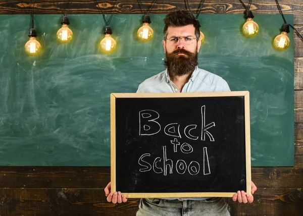 Вчитель в окулярах тримає дошку з написом назад до школи. Чоловік з бородою і вусами на суворому обличчі вітає студентів, креслярські дошки на фоні. Повернутися до концепції школи — стокове фото