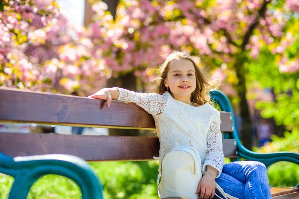 ファッション アクセサリーのコンセプトです。背景には、デフォーカスのさくらのベンチに座っている笑顔に女の子。バックパックでかわいい子では、日当たりの良い春の日をお楽しみください。桜の近くの公園を散歩中の女の子 — ストック写真