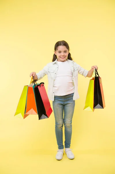 Wenig Helfer. Kind niedlich Fashionista hilft beim Tragen von Paketen beim Einkaufen. kleine Verkäuferin. Kind Mädchen glücklich lächelnd Gesicht trägt Bündel Pakete gelben Hintergrund. Shopaholic mag Shopping — Stockfoto