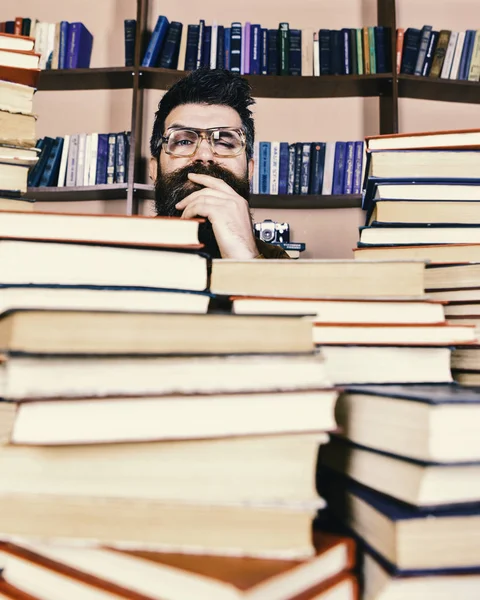 Enseignant ou élève barbu porte des lunettes, s'assoit à table avec des livres, déconcentré. Homme au visage réfléchi entre des piles de livres à la bibliothèque, étagères sur fond. Concept de recherche scientifique — Photo