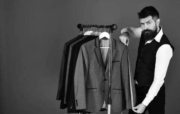 Moderne garderobe keuze concept. Kleermaker met vertrouwen gezicht houdt meetlint in de buurt van aangepaste jassen — Stockfoto