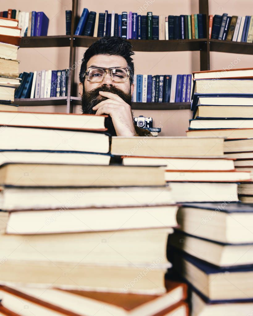 Teacher or student with beard wears eyeglasses, sits at table with books, defocused. Man on thoughtful face between piles of books in library, bookshelves on background. Scientific research concept