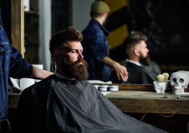 Siyah pelerin ile kaplı sakallı salonların sandalye, ayna arka plan oturur. Hipster berber sakal müşterilerinden olan adam. Berber kavramı. Sakallı hippi Kuaför ve saç kesimi için bekler.