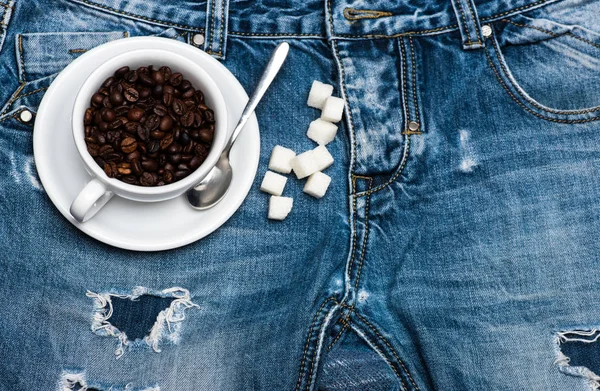 Чашка с кофейными зёрнами, ложкой и рафинированным сахаром лежала на джинсах возле молнии, на джинсовом фоне. Концепция свежего кофе. Кружка полная кофейных зерен на джинсах, вид сверху — стоковое фото