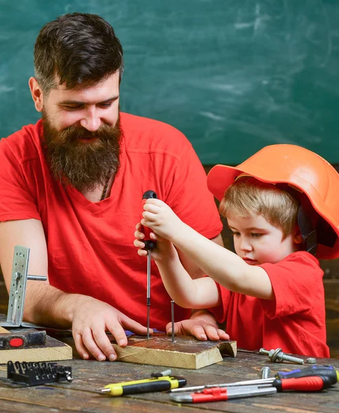 Командная работа и концепция помощи. Отец, родитель с бородой учит маленького сына использовать отвёртку. Мальчик, ребенок занят в защитном шлеме, учится пользоваться отвёрткой с папой. — стоковое фото