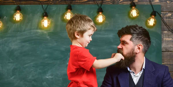 Teacher with beard, father and little son having fun in classroom, chalkboard on background. Child cheerful play with beard of teacher. Fatherhood concept. Dad with beard spend time with son — Stock Photo, Image