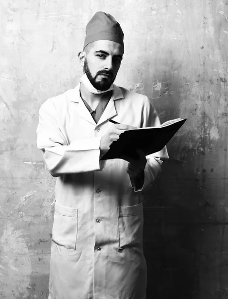 Concept of medicine and professionalism. Surgeon with dark beard in white coat writes in notebook — Stock Photo, Image