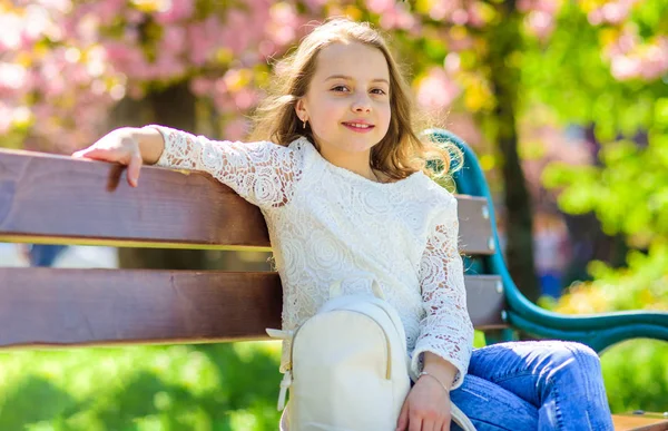 Mode accessoires concept. Meisje op het glimlachen gezicht zit op de Bank, sakura op achtergrond, intreepupil. Meisje ontspannen terwijl wandeling in park in de buurt van kersenbloesem. Schattig kind met rugzak genieten van zonnige lente — Stockfoto