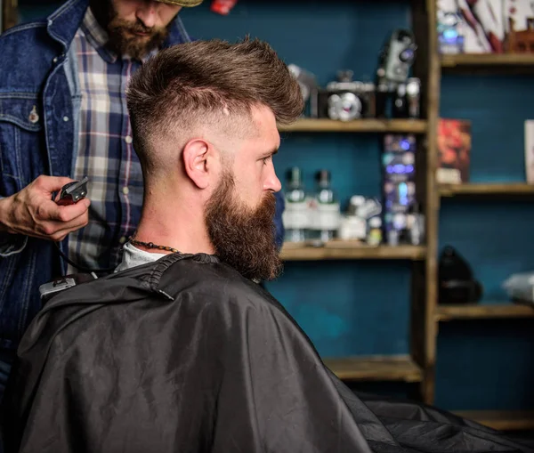 Hairstyle service concept. Hipster bearded client got hairstyle. Barber with trimmer or clipper shaved neck of client. Barber with clipper works on hairstyle for bearded man, barbershop background