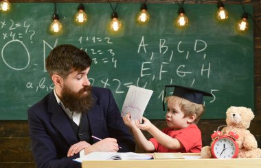 Teacher and pupil in mortarboard, chalkboard on background. Father teaches son elementary knowledge, discuss, explain. Elementary education concept. Kid studies with teacher, listening with attention. clipart