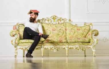 Maço kafasına, çatı gibi açık kitap ile oturuyor. Sakal ve bıyık oturur kanepe, beyaz duvar arka plan üzerinde olan adam. Overstudy kavramı. Adam overdid eğitim ile eski sıkıcı kitap okuma bıkmış.