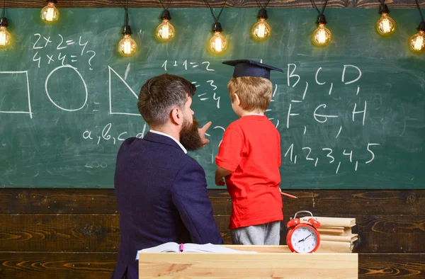 Elev i examen cap lösa ekvation med hjälp av läraren. Man undervisning lilla kid aritmetiska medan pekar på tavlan med siffror. Vuxen hane och en pojke vände tillbaka i klassrummet. — Stockfoto