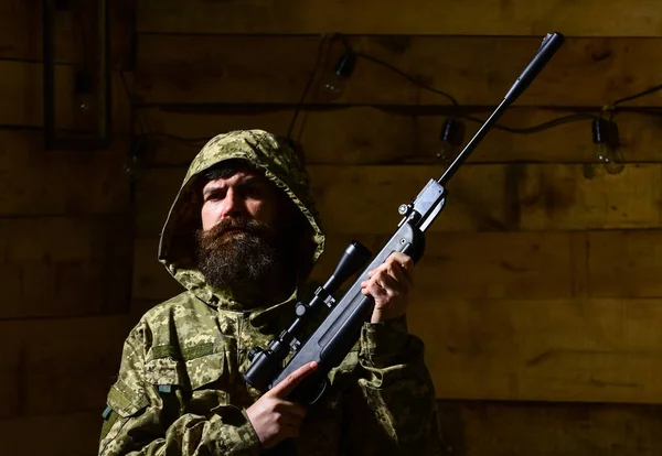 Man with beard wears camouflage hooded clothing, wooden interior background. Macho on strict face at gamekeepers house. Hunter, brutal hipster with gun prepare rifle for hunting. Huntsman concept. — Stock Photo, Image