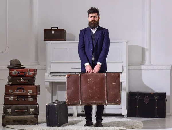 Equipaje y concepto de viaje. Hombre, viajero con barba y bigote con equipaje, lujoso fondo interior blanco. Macho elegante en puestos de cara estrictos y lleva gran maleta vintage . — Foto de Stock