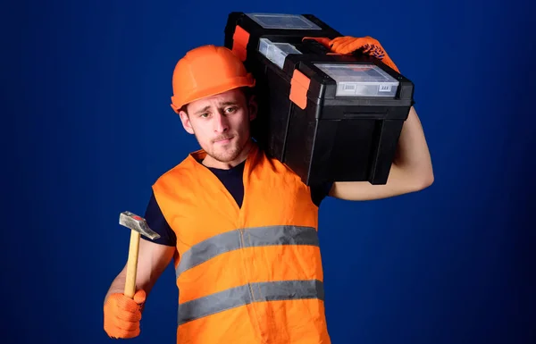 Mann mit Helm, Hut trägt Werkzeugkiste und hält Hammer, blauer Hintergrund. Arbeiter, Reparateur, Reparateur, Bauarbeiter mit ruhigem Gesicht, Handwerker mit Hammer, Werkzeugkiste auf der Schulter. Heimwerkerkonzept. — Stockfoto