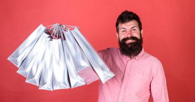 Man with beard and mustache holds shopping bags, red background. Hipster on happy face is shopping addicted or shopaholic. Guy shopping on sales season with discounts. Shopping concept clipart