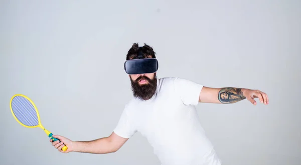 Virtual sport concept. Man with beard in VR glasses beating pitch, grey background. Guy with VR glasses play tennis with racket. Hipster on busy face use modern technology for sport games