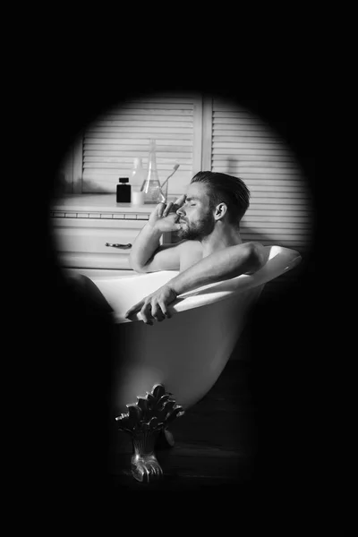 Seksuaalisuuden ja rentoutumisen käsite. Kylpyhuoneessa istuva mies, jolla on kylpytuotteita taustalla. Macho istuu alasti kylpyammeessa vakoillaan avaimenreiästä — kuvapankkivalokuva