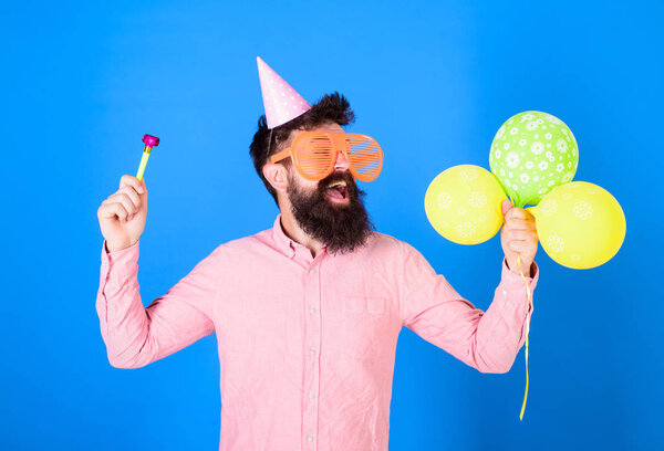 Man with beard and mustache on happy face holds air balloons, blue background. Party concept. Guy in party hat with holiday attributes celebrates. Hipster in giant eyeglasses celebrating birthday