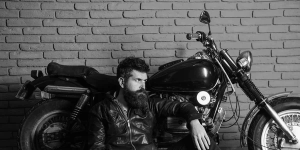 Vahşi motorcu kavramı. Yenilikçi, vahşi Motorcu deri ceketli dalgın yüzündeki motosiklet yakın yerde oturamaz. Sakallı, motorcu motorlu bisiklet garaj, tuğla duvar arka plan yakınındaki deri ceketli adam