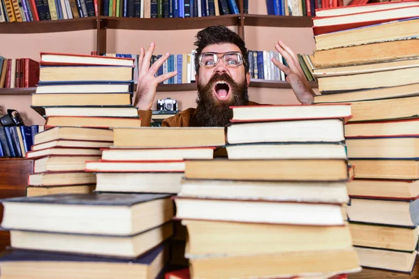 Homme au visage choqué entre des piles de livres à la bibliothèque, étagères sur fond. Enseignant ou élève barbu porte des lunettes, s'assoit à table avec des livres, déconcentré. Concept de découverte scientifique — Photo