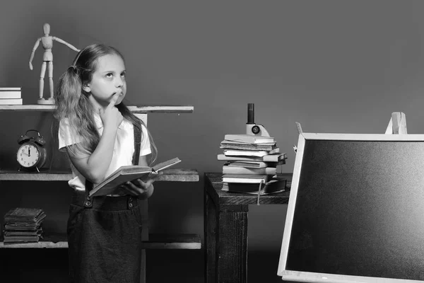 Estudante com cara atenciosa em sua sala de aula. Menina segura livro aberto e fica por quadro-negro em branco — Fotografia de Stock