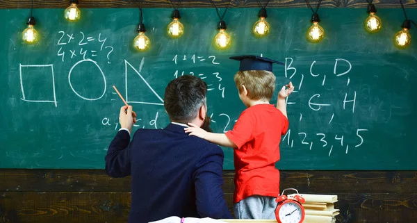 Preparation for school concept. Boy, child in graduate cap listening teacher, chalkboard on background, rear view. Teacher with beard, father teaches little son in classroom, chalkboard on background — Stock Photo, Image
