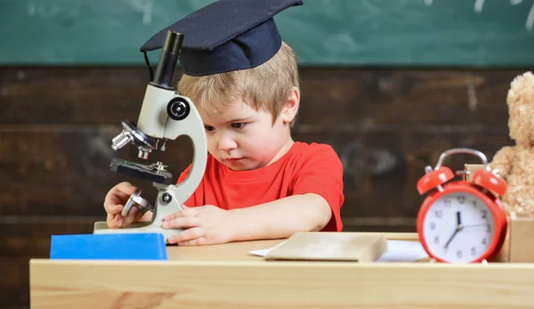 Första tidigare intresserade studera, lärande, utbildning. Kid boy i akademiska cap arbete med mikroskopet i klassrummet, svarta tavlan på bakgrund. Barn på upptagen ansikte nära mikroskopet. Smart unge koncept — Stockfoto