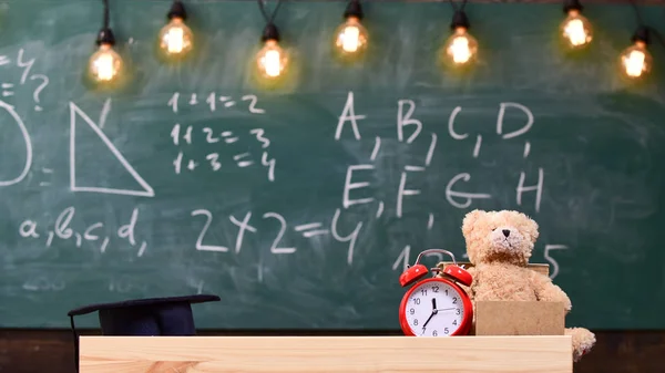 Fechar a sala de aula da escola primária. Sala de aula com quadro-negro no fundo. Mesa infantil com despertador e ursinho de pelúcia. Conceito da escola primária. Sala de aula com mesa com tampa acadêmica em — Fotografia de Stock