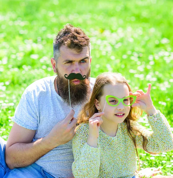 Father posing with mustache and child posing with eyeglasses photo booth attribute. Dad and daughter sits on grassplot, grass on background. Detectives concept. Family spend leisure outdoors — Stock Photo, Image