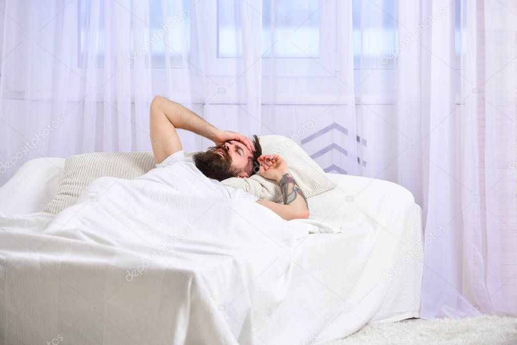 Man in shirt laying on bed awake, white curtain on background. Guy on disappointed painful face waking up in morning. Macho with beard and mustache overslept waking up call. Hangover concept