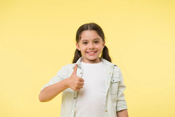 Boldog lány mutogatja a hüvelykujját a sárga háttérben. Kis gyermek mosoly kézmozdulattal. Alkalmi megjelenés. Szépség és divat. Ez tetszik. — Stock Fotó
