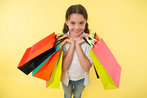 Expert en mode. Enfant expert du shopping mignon aide à transporter des paquets pendant les achats. Petit expert du magasin. Fille shopaholic aime faire du shopping. Enfant fille heureux visage porte paquets tas fond jaune — Photo