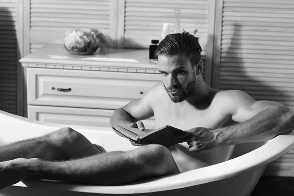 Tipo en el baño con artículos de tocador en el fondo. Hombre con barba y cara sospechosa leyendo libro. Macho sentado desnudo en la bañera, enfoque selectivo . —  Fotos de Stock