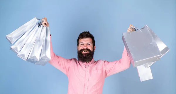 Hombre de camisa rosa con cara feliz y barba larga sosteniendo bolsas de regalo, fiesta de cumpleaños, concepto de celebración. Hipster con barba de moda aislada sobre fondo azul. Barbudo hombre haciendo compras de vacaciones —  Fotos de Stock