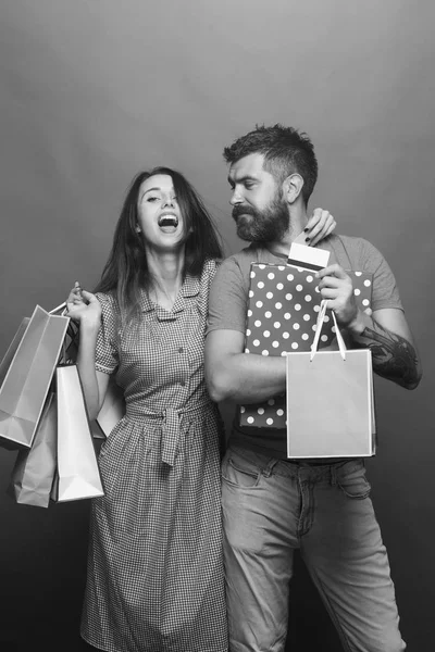 Shopping and fashion concept. Guy with beard and pretty lady with happy face do shopping. Bearded man holds credit cards. Couple in love hugs holding big box and pink shopping bags on green background