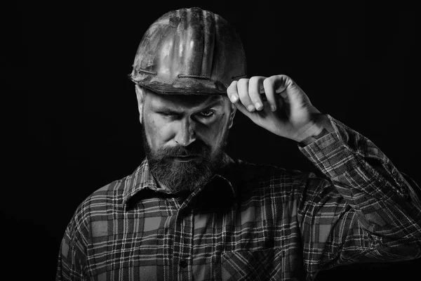 Worker with brutal image wears dirty red helmet and checks it. Builder or miner with thick beard. Construction and mining concept