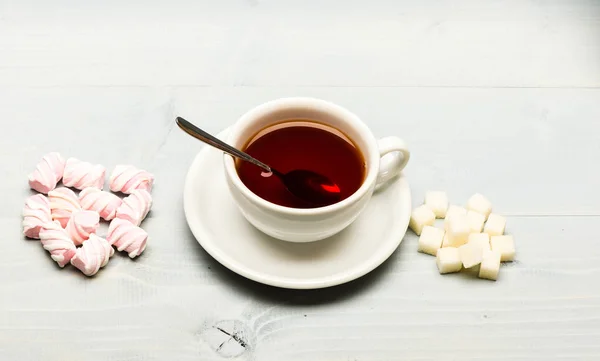 Dryck och sötsaker förberett som efterrätt. Te paus koncept. Mugg med svart te och godis. Mugg fylld med svart bryggt te, sked och högen av marshmallow och raffinerat socker på vit trä bakgrund — Stockfoto