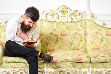 Adam zevk ile eski kitap okuma. Mizahi edebiyat kavramı. Maço üzerinde yüz kitap okuma gülüyor. Barok tarzı kanepe, tutar kitap, beyaz duvar arka plan sakal ve bıyık oturur adam