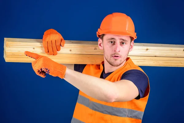 Carpintero, carpintero, fuerte constructor en la cara estricta lleva la viga de madera en el hombro. Concepto de materiales de madera. Hombre en casco, sombrero duro y guantes protectores apuntando dirección, fondo azul —  Fotos de Stock