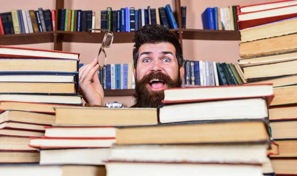 Homem no rosto feliz entre pilhas de livros na biblioteca, estantes de livros no fundo. Conceito de investigação científica. Professor ou estudante com barba usa óculos, senta-se à mesa com livros, desfocado — Fotografia de Stock