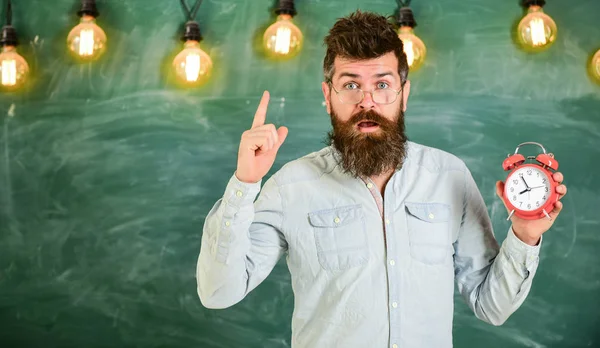 O professor de óculos tem despertador. O hipster barbudo segura relógio, avisando com o dedo indicador, copia espaço. Conceito de disciplina. Homem com barba e bigode no rosto surpreso ficar em sala de aula — Fotografia de Stock