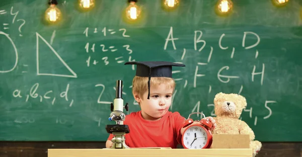 Skolan paus koncept. Barn på koncentrerade ansikte ser väckarklocka. Elev väntar skolan paus. Kid boy i akademiska cap nära Mikroskop, klocka och leksak i klassrummet, svarta tavlan på bakgrund — Stockfoto