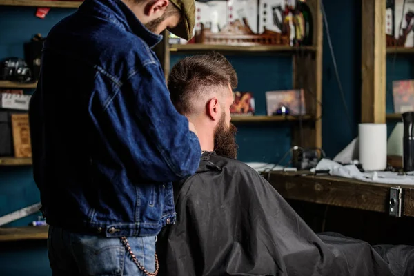 Hairstyle service concept. Barber with clipper works on hairstyle for bearded man, barbershop background. Hipster bearded client got hairstyle. Barber with trimmer or clipper shaved neck of client