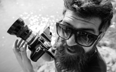 Arka plan su yüzeyine giyer gözlüklü, sakal ve bıyıklı adam. Hipster gülümseyen yüzü üzerinde eski moda kamera tutar. Turist fotoğrafçı kavramı. Doğa nehir ya da göl yakınındaki çekim adam