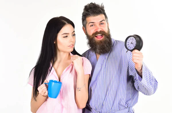 Perfect morning concept. Couple, family woke up on time. Couple in love, young family in pajama, bathrobe stand isolated on white background. Girl with coffee cup, man holds clock in hand