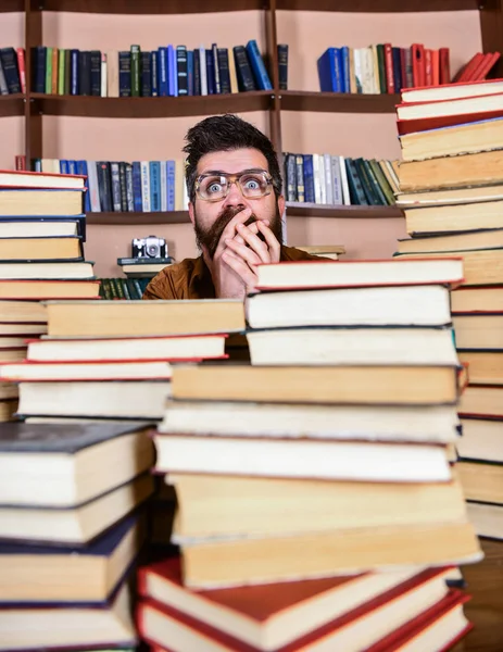 Homme au visage choqué entre des piles de livres à la bibliothèque, étagères sur fond. Concept de découverte scientifique. Enseignant ou élève barbu porte des lunettes, s'assoit à table avec des livres, déconcentré — Photo